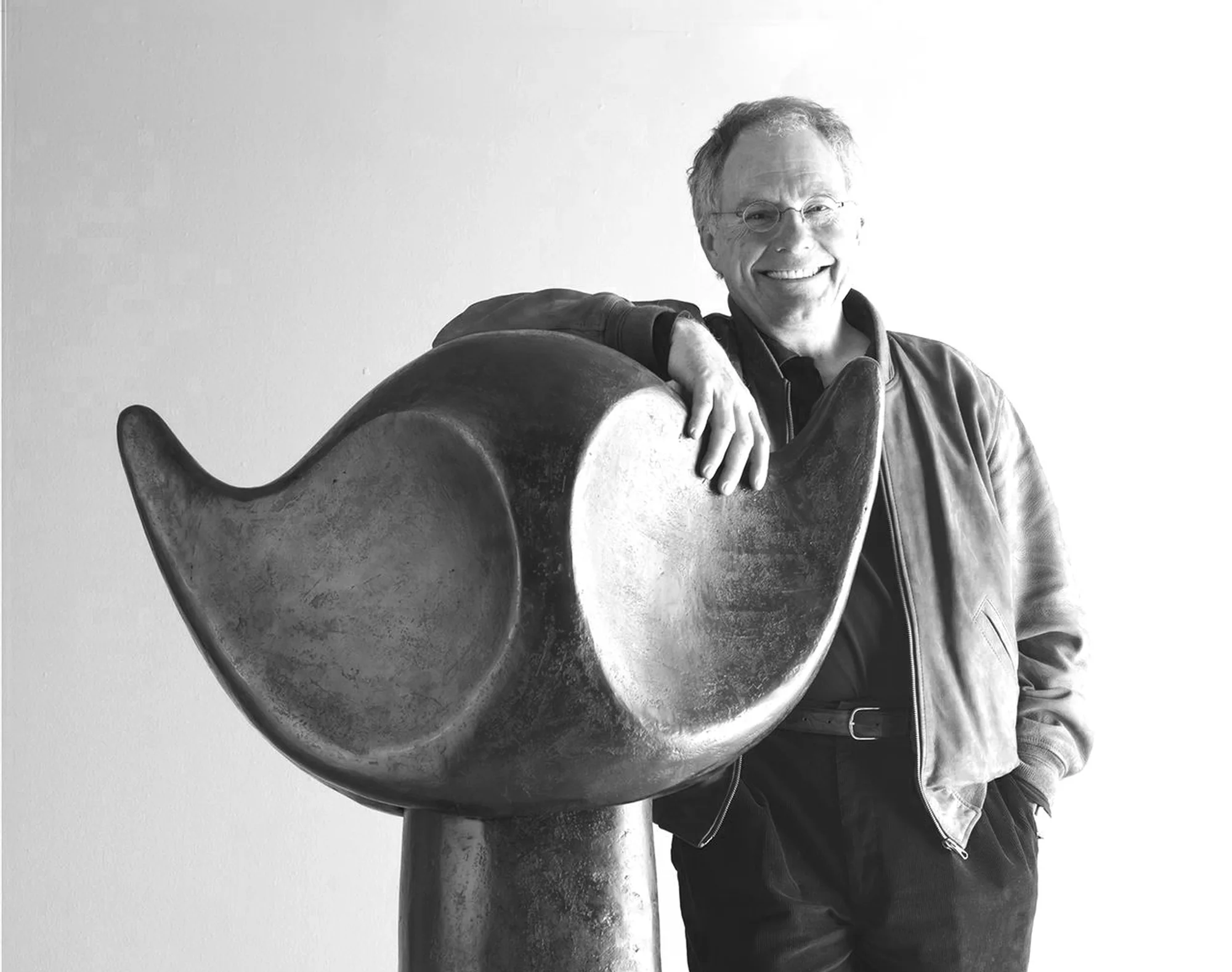 Bildhauer Werner Pfändler lächelnd mit der Hand auf einer großen geschwungenen Skulptur, Schwarz-Weiß-Aufnahme