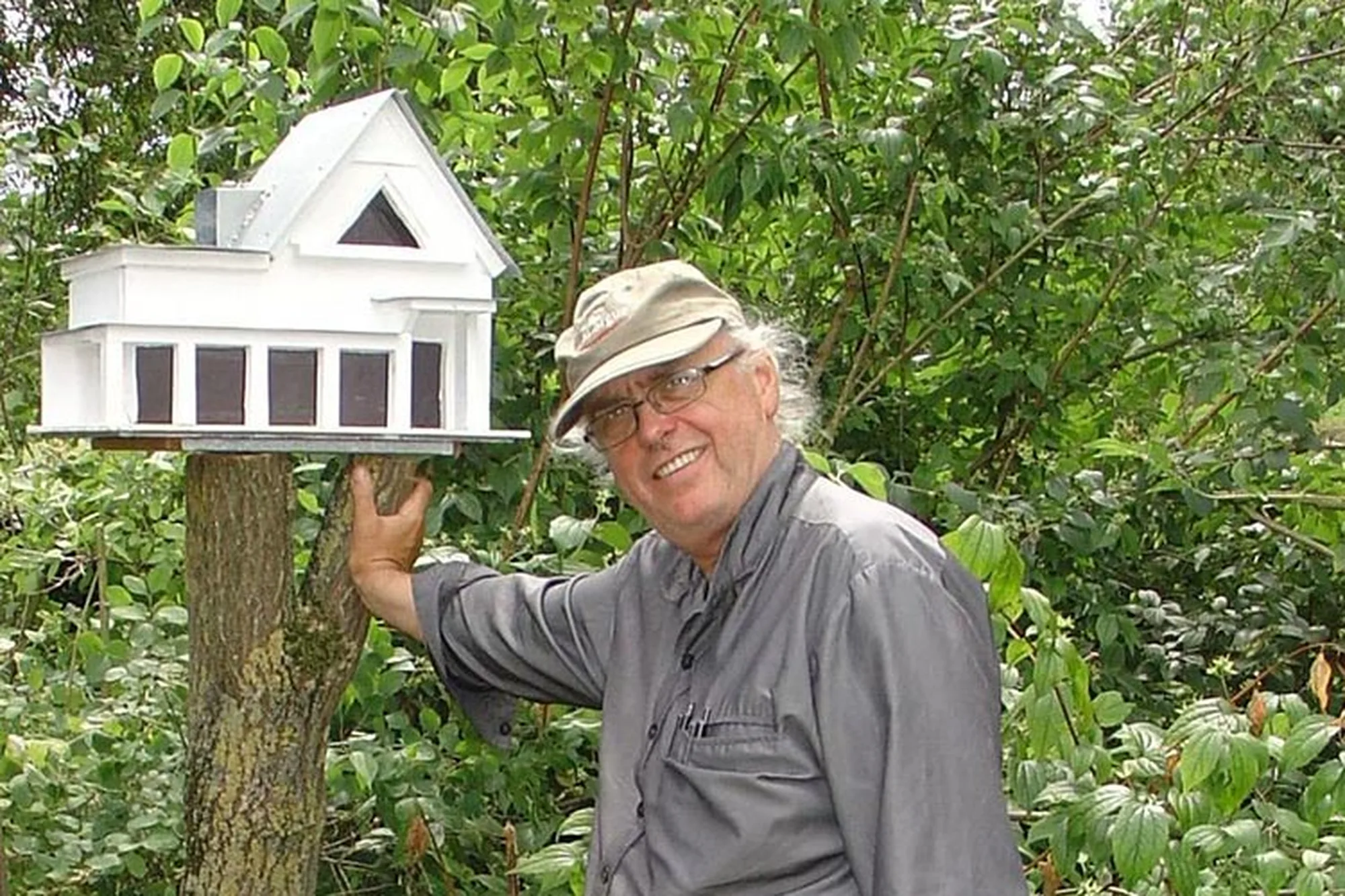 Bildhauer Raimund Schmelter mit Kappe im Garten neben einem weißen Vogelhaus in Villenform