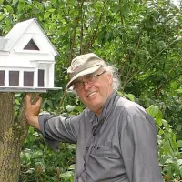 Bildhauer Raimund Schmelter mit Kappe im Garten neben einem weißen Vogelhaus in Villenform